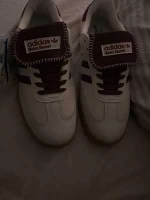 Adidas Wales Bonner sneakers vita/lila - Unika Adidas Wales Bonner sneakers i vitt och lila med klassiska ränder på sidan och grov sula i gummi. Tungan har Wales Bonner-logga och dekorativ söm. Skorna har låga skaft och är tillverkade i läder och textil för en retrovibe.