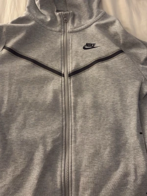 Grå zip hoodie från Nike XL-158-170 - Snygg grå hoodie från Nike i storlek XL med dragkedja framtill och svart Nike-logga på bröstet. Hoodien har en svart detalj över bröstet och huva. Tillverkad i mjukt bomullsmaterial, perfekt för chill dagar eller träning. Två små små rivningar lägst ned men som knappt syns.