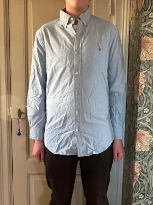 Ljusblå skjorta från Ralph Lauren - Klassisk ljusblå skjorta från Ralph Lauren med button-down krage och broderad logga på bröstet. Skjortan har långa ärmar, normal passform och är tillverkad i bomull. Perfekt till jeans eller chinos för en clean look.