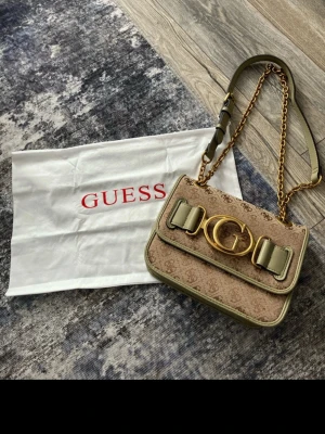 Beige axelväska från Guess - Snygg beige axelväska från Guess med monogrammönster, guldfärgad kedja och stort G-spänne framtill. Väskan har detaljer i ljusgrönt och justerbar axelrem i både läder och metall. Perfekt för dig som vill ha en trendig och stilren accessoar.
