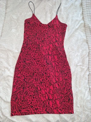 Röd leopardmönstrad klänning - Snygg röd kortklänning med tunna axelband och leopardmönster i svart. Klänningen har en figurnära passform och är perfekt för dig som vill sticka ut. Materialet känns mjukt och följsamt, troligen polyester eller liknande. Använd fåtal gånger💥