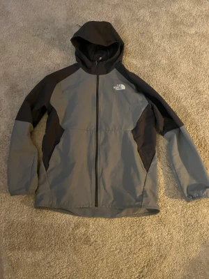 Grå och svart vindjacka The North Face - Snygg vindjacka från The North Face i grått och svart med huva och dragkedja framtill. Jackan har två sidofickor och logga på bröstet. Tillverkad i lätt och slitstarkt material, perfekt för blåsiga dagar. Storlek XXl för barn men sitter som S