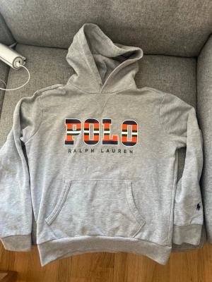 Polo Ralph Lauren Hoodie - Snygg grå hoodie från Polo Ralph Lauren med stor färgglad POLO-logga på bröstet och broderad logga på ärmen. Klassisk känguruficka framtill och ribbade muddar. Tillverkad i mjuk bomullsblandning och har en stor huva för extra chill vibe. Pris kan diskuteras vid snabba affär😇