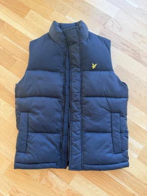 Svart dunväst från Lyle & Scott - Snygg Svart dunväst från Lyle & Scott med gul broderad logga på bröstet. Västen har hög krage, dragkedja och tryckknappar framtill samt två stora fickor. Perfekt för lager på lager och streetwear-stil.