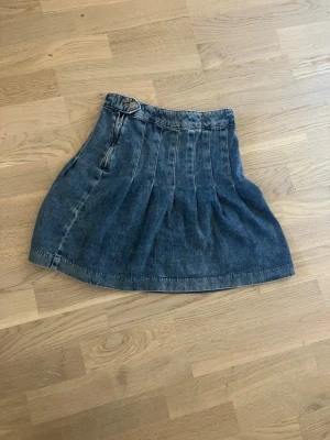 Blå jeanskjol med dragkedja - Säljer en blå jeanskjol i kort modell med veckad design. Kjolen har en synlig dragkedja och knapp i midjan för en cool detalj. Perfekt till sneakers eller boots för en avslappnad look.