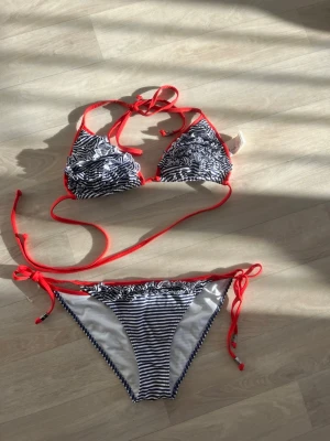 Randig bikini med röda knytband - Snygg bikini med svartvita ränder och röda knytband. Överdelen har volangdetaljer och klassisk triangelmodell. Underdelen har knyt i sidorna för justerbar passform. Perfekt för stranden eller poolen!