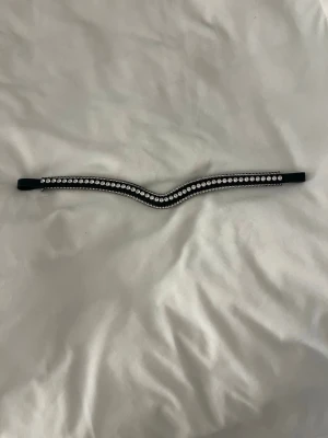 Svart pannband med strassdetaljer - Snyggt svart pannband med en V-formad design, dekorerat med två rader glittrande strass. Perfekt för att ge din frisyr extra bling och stil. Materialet är troligtvis konstläder eller textil med infällda stenar. Passar till dig som vill sticka ut lite extra.