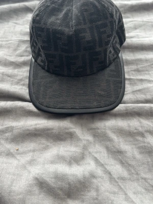 Svart keps med FF-mönster från Fendi - Säljer en svart keps från Fendi med det klassiska FF-monogrammet över hela kepsen. Kepsen har böjd skärm med svart kant och justerbart band bak. Materialet är tyg med exklusiv känsla och insidan är fodrad för extra komfort.
