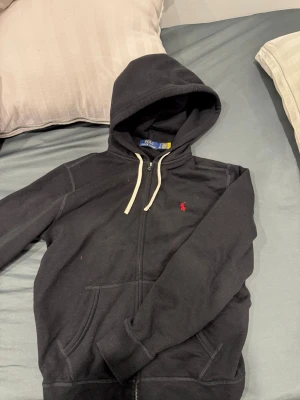 Svart hoodie från Polo Ralph Lauren - Svart hoodie från Polo Ralph Lauren med dragkedja, vit dragsko i huvan och den klassiska röda loggan broderad på bröstet. Tillverkad i mjukt bomullsmaterial och har kängurufickor framtill. Perfekt för en avslappnad och stilren look.