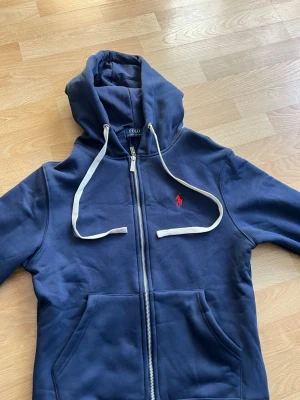Ralph lauren zip hoodie - Snygg marinblå hoodie från Polo Ralph Lauren med dragkedja inte anävnd en ändå gång, var lite för liten så säljer den direkt istället, vit dragsko och klassiska röda loggan broderad på bröstet. Hoodien har känguruficka och är tillverkad i mjukt bomullsmaterial. Perfekt för en chill och stilren look.