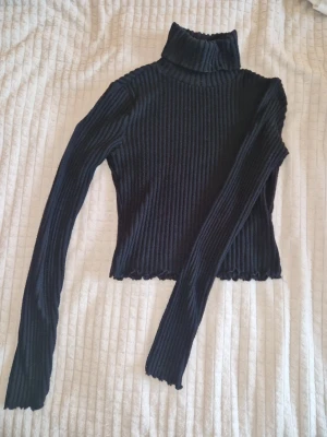 Svart ribbad polotröja gina tricot  - Säljer en svart ribbad,kortare polotröja med lång ärm och hög krage. Tröjan är figurnära och har vågig kant nedtill, och ärmslut💥Materialet är 95% polyester och 5% elastan vilket gör den stretchig och bekväm.