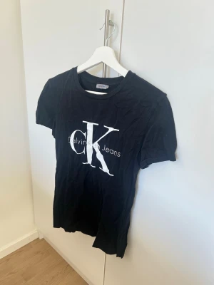 Svart Calvin Klein t-shirt - Snygg svart t-shirt från Calvin Klein med stort vitt CK Jeans-tryck på bröstet. Klassisk rund hals och korta ärmar. Perfekt till jeans eller shorts för en clean och enkel look.