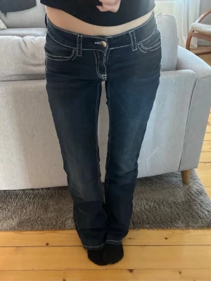 Mörkblå bootcut jeans med kontrastsömmar - Säljer ett par mörkblå bootcut jeans med vita kontrastsömmar och låg midja. Snygg paljett detalj på fickorna. Gina Young storlek 164. 36cm över midjan och 77cm innerbenslängd 