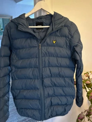 Blå pufferjacka från Lyle & Scott - Snygg blå pufferjacka från Lyle & Scott med huva och dragkedja framtill. Jackan har det klassiska gula logomärket på bröstet och är quiltad för extra värme. Perfekt för kalla dagar och har en modern, avslappnad passform. Jag e 170 centimeter lång och den passar perfekt.