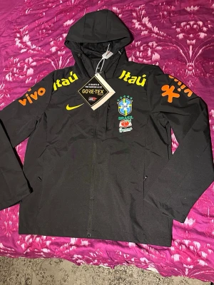 Svart Brasil  vindjacka GORE-TEX i L och XL -  XL passar L/M. Snygg svart vindjacka från Nike med Brasilien CBF-logga och sponsormärken som Itaú, Vivo och Guaraná. Jackan har huva, dragkedja framtill och är tillverkad i slitstarkt GORE-TEX-material. Perfekt för dig som gillar sportig stil och vill visa stöd för Brasilien.