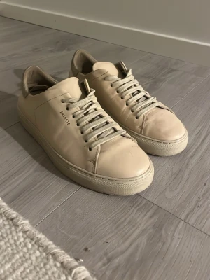 Beige sneakers från Axel Arigato - Stilrena beige sneakers från Axel Arigato i läder med matchande snörning och diskret logga på sidan. Klassisk låg modell med vit sula och mjuk mockadetalj på hälen. Perfekta för dig som gillar minimalistisk och clean stil.
