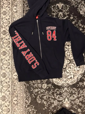  Superdry hoodie - Snygg mörkblå hoodie från Superdry med dragkedja, huva och fickor. Stor röd och vit text 'SUPER ATHL.' på ärmen och 'SUPERDRY 84' på bröstet. Tillverkad i mjukt bomullsmaterial, perfekt för en sportig och avslappnad stil.