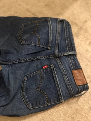 Mörkblå Levi's jeans med raka ben - Klassiska mörkblå jeans från Levi's med raka ben och snygga kontrastsömmar. Jeansen har två bakfickor med den ikoniska V-formade sömmen och en brun läderpatch i midjan. Tillverkade i slitstark denim med en tidlös look.