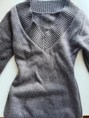 Grå stickad tröja med v-ringning - Mysig grå stickad tröja med bred ribbad v-ringning och långa ärmar. Tröjan har en figurnära passform och ribbade muddar vid ärmslut och nederkant. Perfekt för lager på lager och enkel att matcha med olika stilar.
