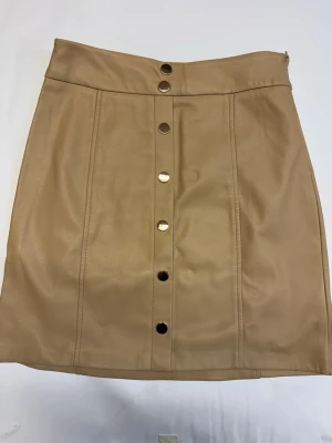 Beige kjol i skinnimitation med knappar - Snygg beige kortkjol i skinnimitation med dekorativa metallknappar framtill. Kjolen har hög midja och raka sömmar som ger en clean look. Knappt använd. 