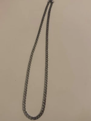 Silverfärgat kedjehalsband - Stilrent halsband i silverfärg med platt, bred kedja. Halsbandet har en klassisk design med tätt sammanlänkade länkar som ger ett snyggt och modernt intryck. Perfekt för dig som gillar minimalistiska accessoarer. Materialet ser ut att vara metall.