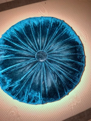 Rund sammetskudde i petrolblå - Säljer en rund kudde i mjuk petrolblå sammet med dekorativa veck och en knapp i mitten. Kudden har en lyxig känsla och passar perfekt som inredningsdetalj i soffan eller på sängen.