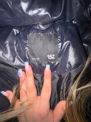 Svart pufferjacka från 157 - Snygg svart pufferjacka från 157 med glansig finish och huva. Jackan har dragkedja framtill och quiltad design som håller värmen. Perfekt för kalla dagar och enkel att matcha med olika outfits.