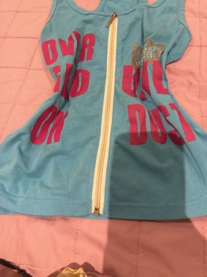 Blå ärmlös topp med dragkedja - Säljer en blå ärmlös topp med dragkedja framtill och stora rosa bokstäver som tryck. Toppen har en glittrig detalj på bröstet och är i ett mjukt bomullsmaterial. Perfekt för dig som gillar sportig och färgglad stil!