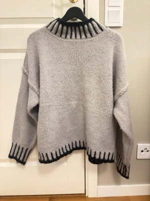 Grå stickad tröja från Flex Moda - Mysig grå stickad tröja från Flex Moda med bred polokrage och svarta detaljer vid hals, ärmslut och nederkant. Oversized passform och ribbstickade muddar ger en avslappnad look. Perfekt för kyliga dagar. Storlek M. Köpt i Spanien! 
