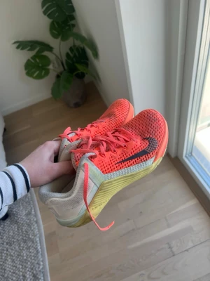 Nike Metcon orange mesh  - Säljer ett par snygga Nike Metcon i orange mesh med svart swoosh på sidan och gula detaljer på sulan. Skorna har lågt skaft och snörning framtill. Perfekta för dig som gillar sportig och färgstark stil.