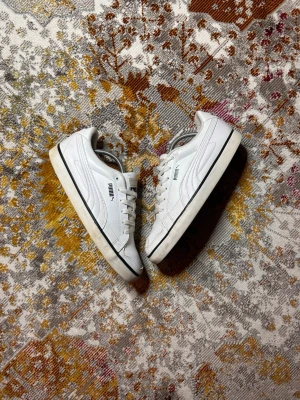 Vita Puma sneakers i läder - Snygga vita sneakers från Puma i klassisk låg modell. Skorna är tillverkade i läder med svarta detaljer och har den ikoniska Puma-loggan på sidan. Perfekta för en clean och stilren look.