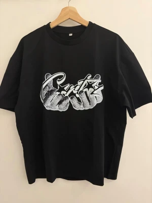 Corteiz t-shirt med grafiskt tryck - Cool svart t-shirt med ett stort vitt grafiskt tryck framtill som föreställer två händer och text i streetstil. T-shirten har rund hals och korta ärmar. Perfekt för dig som gillar streetwear och vill sticka ut.