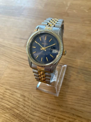 Shivas Quartz A2524 37mm Armbandsur - Shivas Quartz armbandsur med ett tvåfärgat länkarmband i rostfritt stål. Klockan har en blå urtavla med guldfärgade visare och indexmarkeringar. Datumvisning finns vid klockan 3. Boetten är i silvertonat stål med en räfflad guldfärgad bezel. Baksidan av boetten är märkt "STAINLESS STEEL CASE BACK A2524 DUST PROOF HIGH QUALITY". Klockan är i fungerande men använt skikc. Urverket går att ställa in men urtavlan är något lös. Behöver nytt batteri.  Begagnat värde: 500 SEK Mitt pris: 289 SEK  Skickas