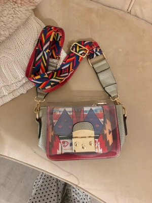 Transparent handväska med färgglatt band - Cool transparent handväska med gråa sidor, guldigt lås och bred axelrem i färgglatt mönster. Inuti finns en liten väska med grafiskt tryck i rött, blått och orange. Perfekt för dig som gillar unika accessoarer och färg. Jag köpte den i Italien så det är inget specifikt märke på den! Den är i helt nytt skick har aldrig använt!💖💖