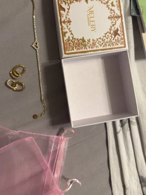 Guldarmband och örhängen med pärlor från Melery - Snyggt guldarmband från Melery London med små gnistrande pärlor längs hela bandet och ett hjärta i mitten. Armbandet har en tunn, elegant kedja och justerbar låsning. Perfekt accessoar för att lyfta din stil. Samt ett par örhängen med samma guldiga stil och pärlor.Aldrig använts.