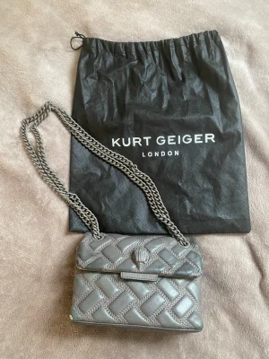 Grå quiltad handväska Kurt Geiger - Snygg grå quiltad handväska från Kurt Geiger med kedjerem och lock med magnetknäppning. Väskan har ett elegant mönster och metallogo framtill. Perfekt storlek för det viktigaste och levereras med original dustbag. Liten defekt i hörnet av väskan men märks inte. 