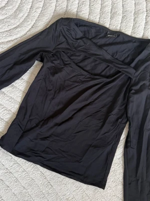 Svart långärmad topp med asymmetrisk hals - Snygg svart långärmad topp med unik asymmetrisk halsringning och smal passform. Tillverkad i mjuk och stretchig bomullsblandning. Perfekt till fester
