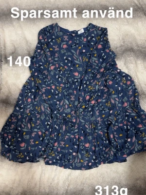 Blommig blå klänning med volang - Söt blå klänning med blommigt mönster. Klänningen har volangdetaljer och lång ärm, perfekt för en lekfull och trendig look.🌸 Sparsamt använd. Storlek 140