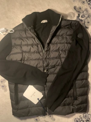 Moncler cardigan  - Snygg svart pufferjacka från Moncler med quiltad front och mjuka stickade ärmar. Jackan har dragkedja framtill, ståkrage och två fickor med dragkedja. Moncler-logga på ärmen ger en exklusiv touch. Perfekt för kalla vinterdagar.