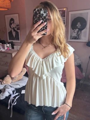 Vit volangtopp med v-ringning - Söt vit topp med v-ringning och stora volangdetaljer framtill. Toppen har korta ärmar och ett flowy, lätt utställd passform. Perfekt att styla med jeans eller kjol för en trendig look.