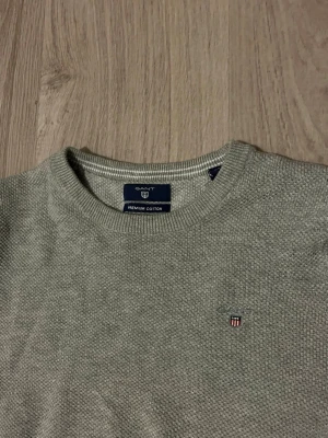 Ljusgrå stickad tröja från Gant - Snygg ljusgrå stickad tröja från Gant i premium bomull. Tröjan har rund halsringning, diskret Gant-logga broderad på bröstet och ribbade muddar vid ärmslut och nederkant. Perfekt för en clean och stilren look. Lowkey grish också