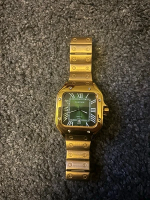 Guldfärgad Santos klocka - Snygg Santos klocka i guldfärgat metallarmband och fyrkantig boett. Klockan har svart urtavla med romerska siffror och datumvisning. Klassisk design med skruvdetaljer på länken och boetten. Perfekt accessoar för dig som gillar exklusiv stil.