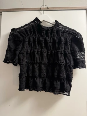 Svart spetsblus med volanger - Trendig svart blus i spets med volangdetaljer och halvtransparent material. Blusen har korta ärmar och hög hals, perfekt för att lyfta vilken outfit som helst. Snygg att styla med både jeans och kjol.