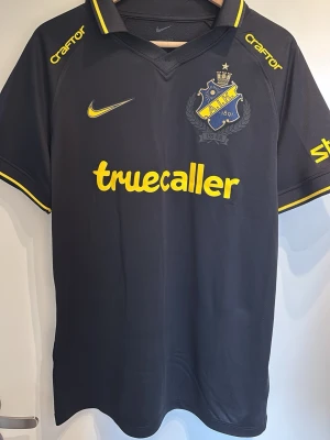 Svart AIK matchtröja 2026 M - Snygg svart AIK matchtröja från Nike med gula detaljer och klubbmärke på bröstet. Tryck med sponsorer som Truecaller och Svea samt nummer 36 och namnet Yohanna på ryggen. Klassisk krage och korta ärmar, perfekt för fotbollsträning eller match.
