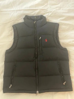 Svart dunväst från Polo Ralph Lauren - Snygg svart dunväst från Polo Ralph Lauren med hög krage, två dragkedjefickor och en bröstficka med röd logga. Västen har quiltad design och stängs med dragkedja. Perfekt för lager-på-lager och streetwear-vibe.