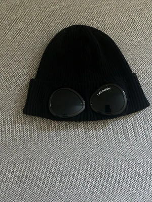 Svart mössa med glasögon från C.P. Company - Säljer min CP Company mössa som endast är använd 1 gång, så den är i princip som ny. Inga defekter eller slitage. 🧢 Färg: Svart 📏 Storlek: One size ✨ Skick: 10/10 Mitt pris: 550kr(kan diskuteras)