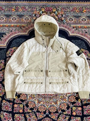 Beige pufferjacka från Stone Island - Säljer en beige pufferjacka från Stone Island med avtagbar huva och ikonisk patch på ärmen. Jackan har quiltad design, två stora fickor framtill med knappar och dragkedja. Perfekt för kalla dagar och riktigt snygg streetstil.