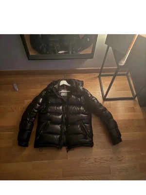 Svart pufferjacka från Moncler - Säljer en svart pufferjacka från Moncler med glansigt yttertyg och klassisk Moncler-logga på ärmen. Priset är prutbart vid en snabbaffär. 