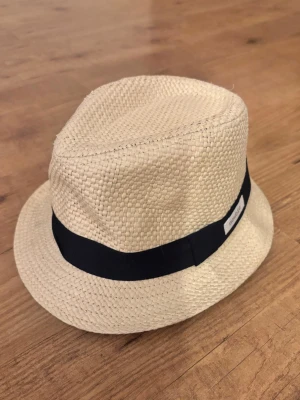 Beige stråhatt från newbie - Snygg beige stråhatt från newbie med klassisk form och ett brett svart band runt kullen. Perfekt accessoar för soliga dagar och ger en chill vibe till din outfit. Tillverkad i flätat stråmaterial som känns lätt och luftigt på huvudet.