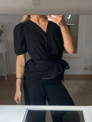 Svart omlottopp med knytband - Snygg svart omlottopp med puffiga korta ärmar och brett knytband i midjan. Toppen är aldrig använd💕